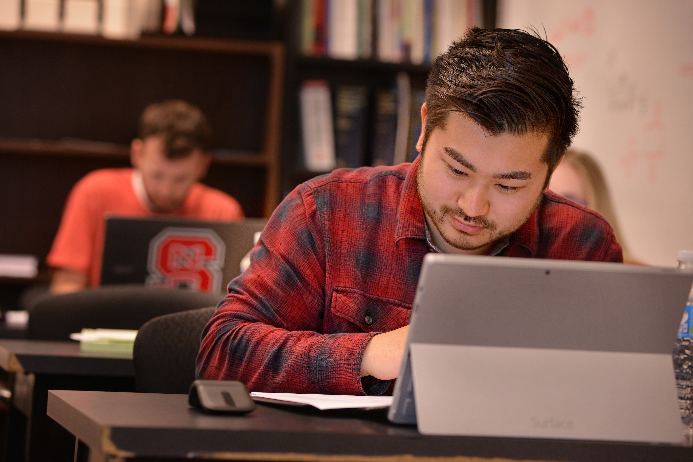 Student at desk with computer and notebook. Books and whiteboard in the background.