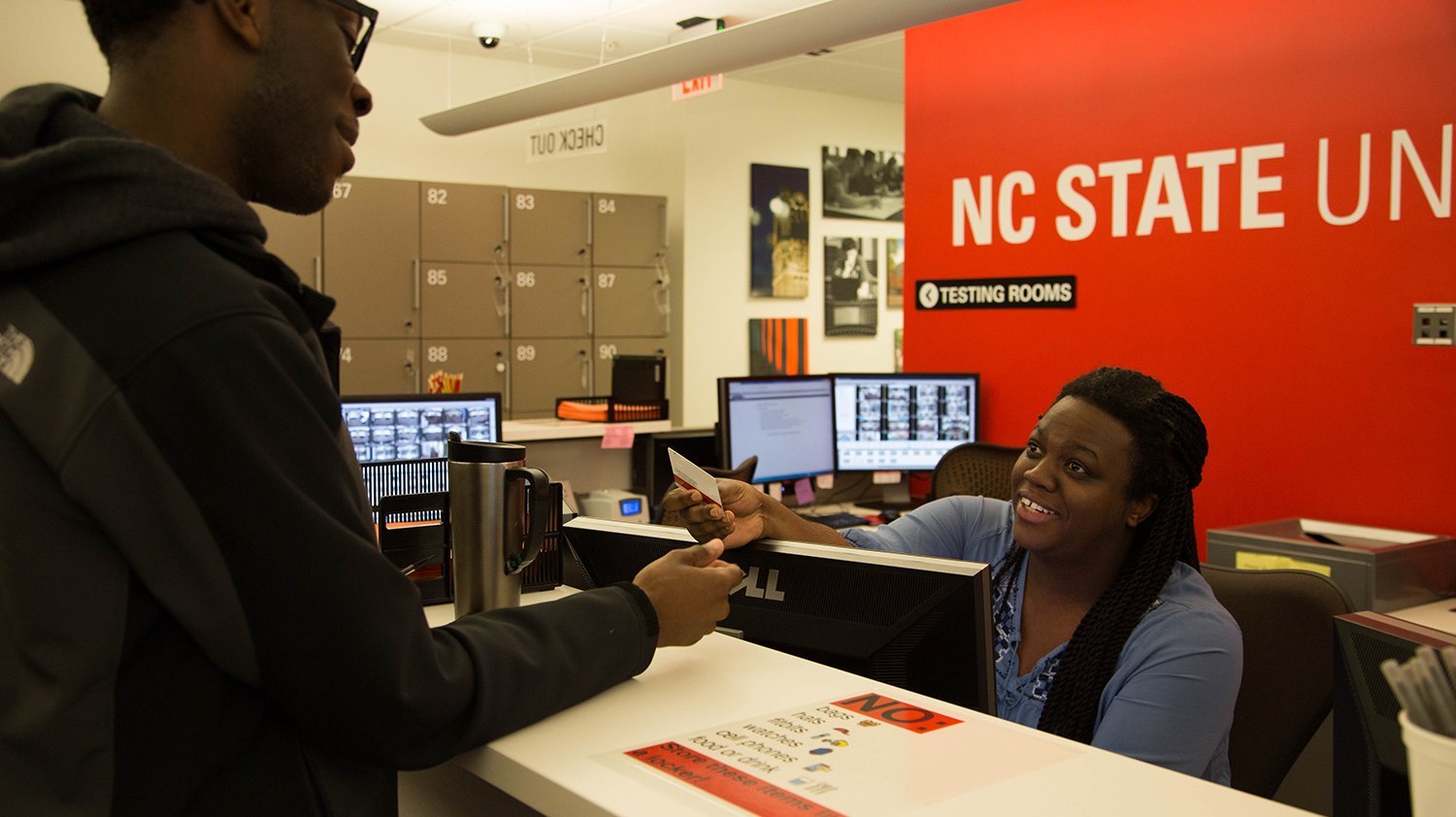 Lead Testing Associate Whitney Hicks (right) checks in a student for an exam at the Centennial Campus Test Center. Whitney is sitting behind a desk and passing the student an ID back after verifying it.