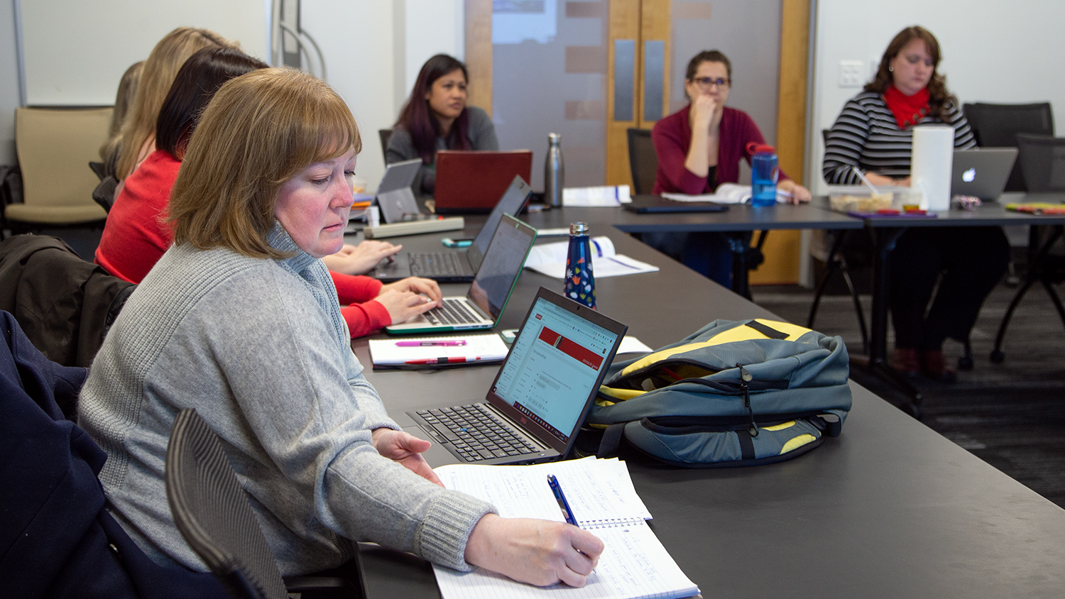Christine Cranford writes down important notes during a face-to-face meeting of the Online Course Improvement Program in spring 2019. Christine is sitting at a table with other people in the distance.