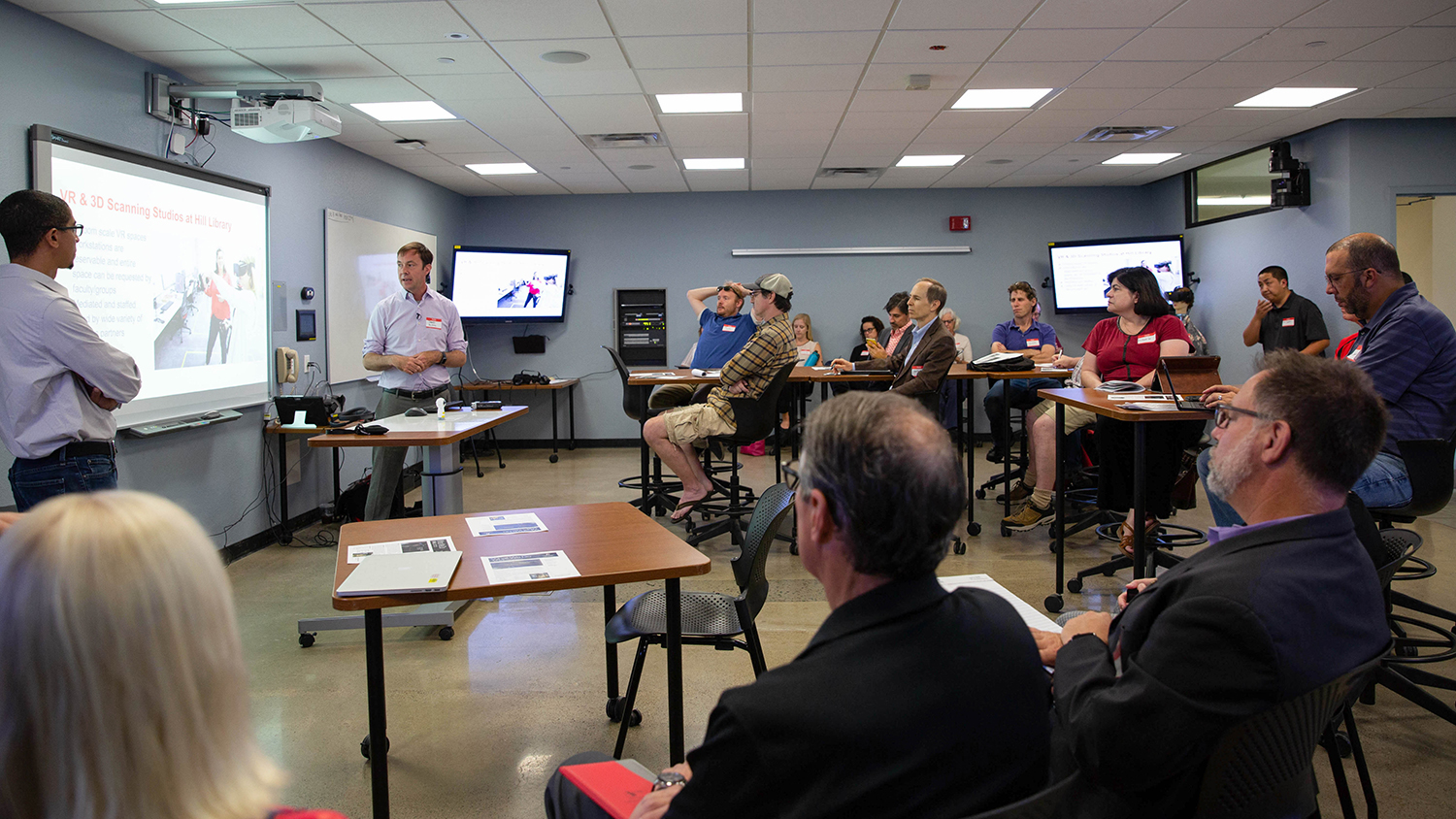 Mike Cuales (front left) and David Woodbury (front right) give a presentation at San Diego State University about immersive learning technologies and spaces. Cuales and Woodbury are at the front of the room and there are participants sitting at tables engaged in their presentation.