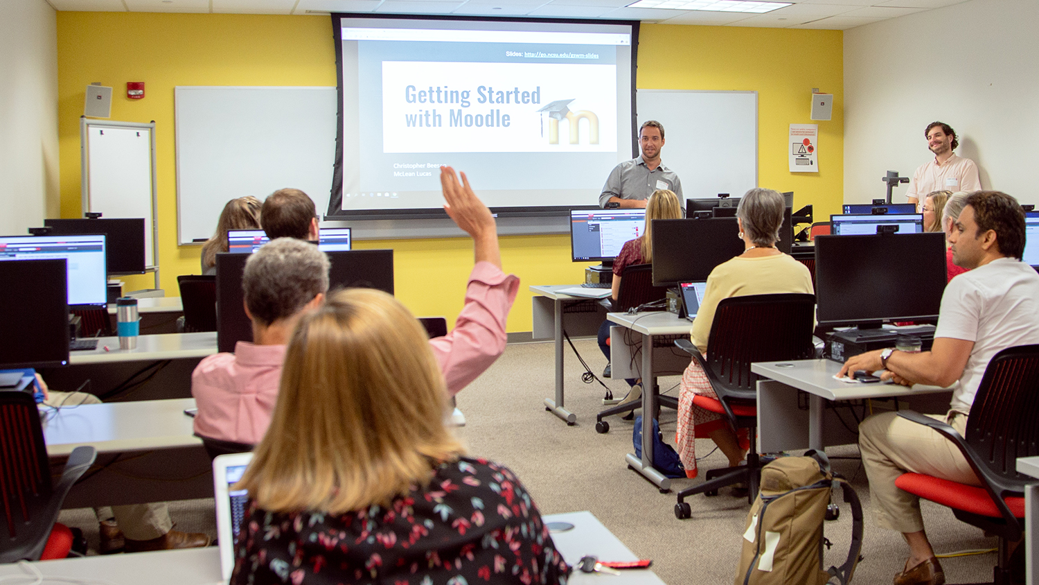 Christopher Beeson and McLean Lucas stand at the front of a computer lab room. There are participants at computer stations around the room. One participant is raising his hand. The screen says "Getting Started with Moodle."
