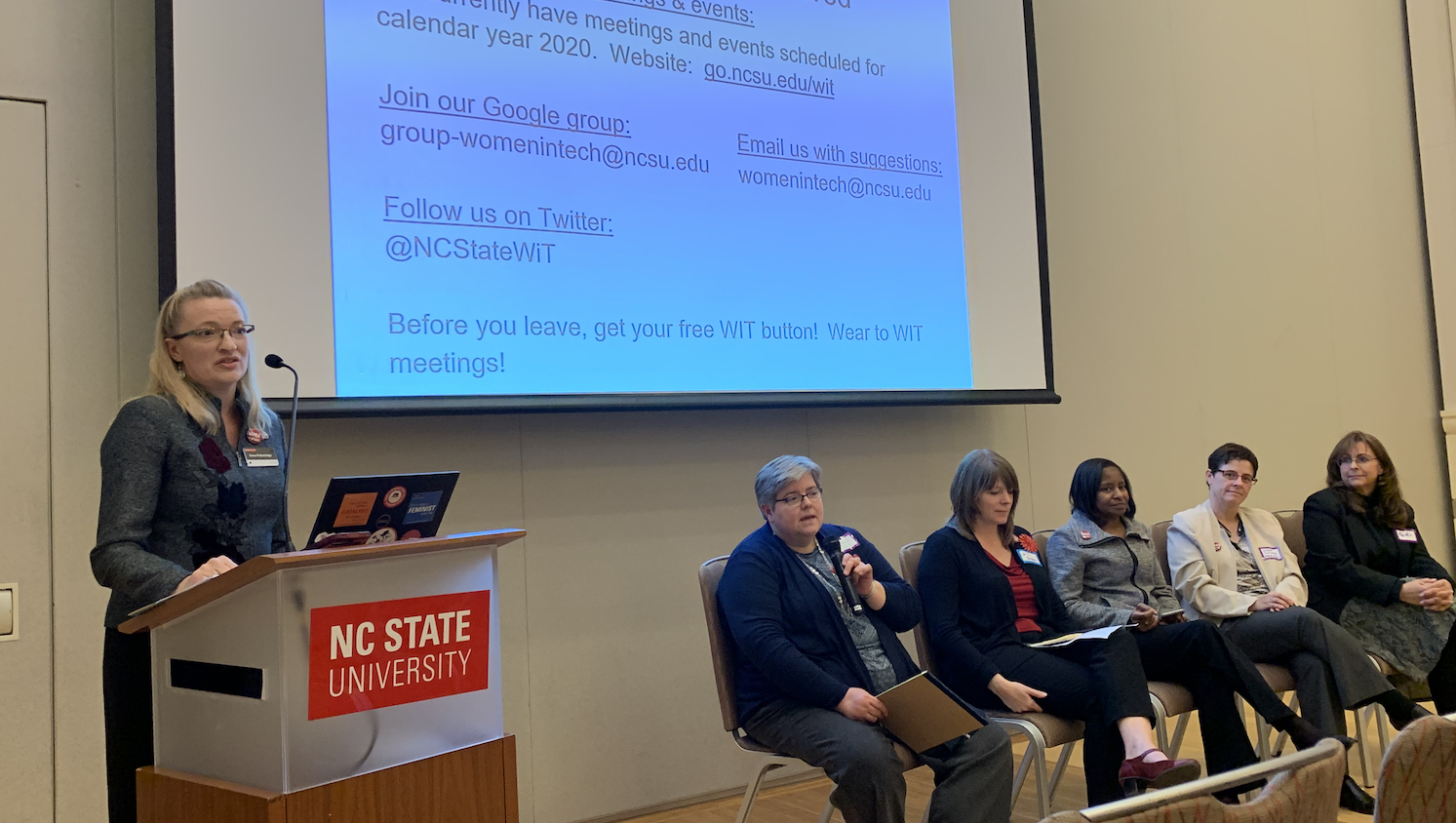 The Women in Technology (WIT) interest group kicked off at the 2019 IT Community Event. Donna Petherbridge (far left) facilitates a leadership panel with other members of the WIT steering committee. Pictured left to right: Susan West, Jill Sexton, Mardecia Bell, Debbie Carraway, and Gwen Hazelhurst. Donna Petherbridge is at a podium and the other members are sitting in chairs at the front of the room.