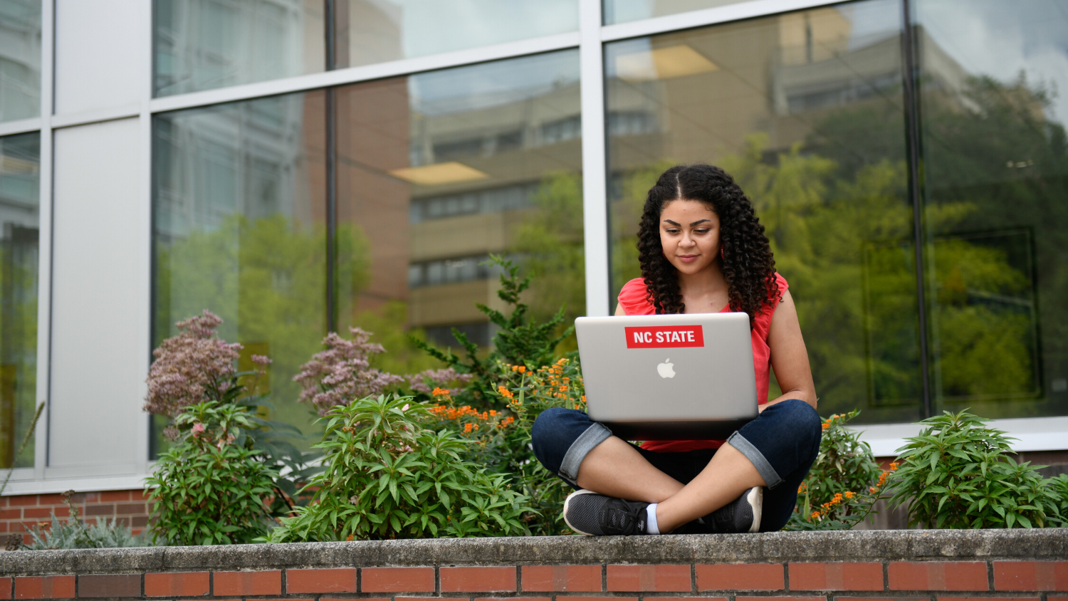 Young woman sitting on a brick ledge with a laptop in front of large windows and greenery.