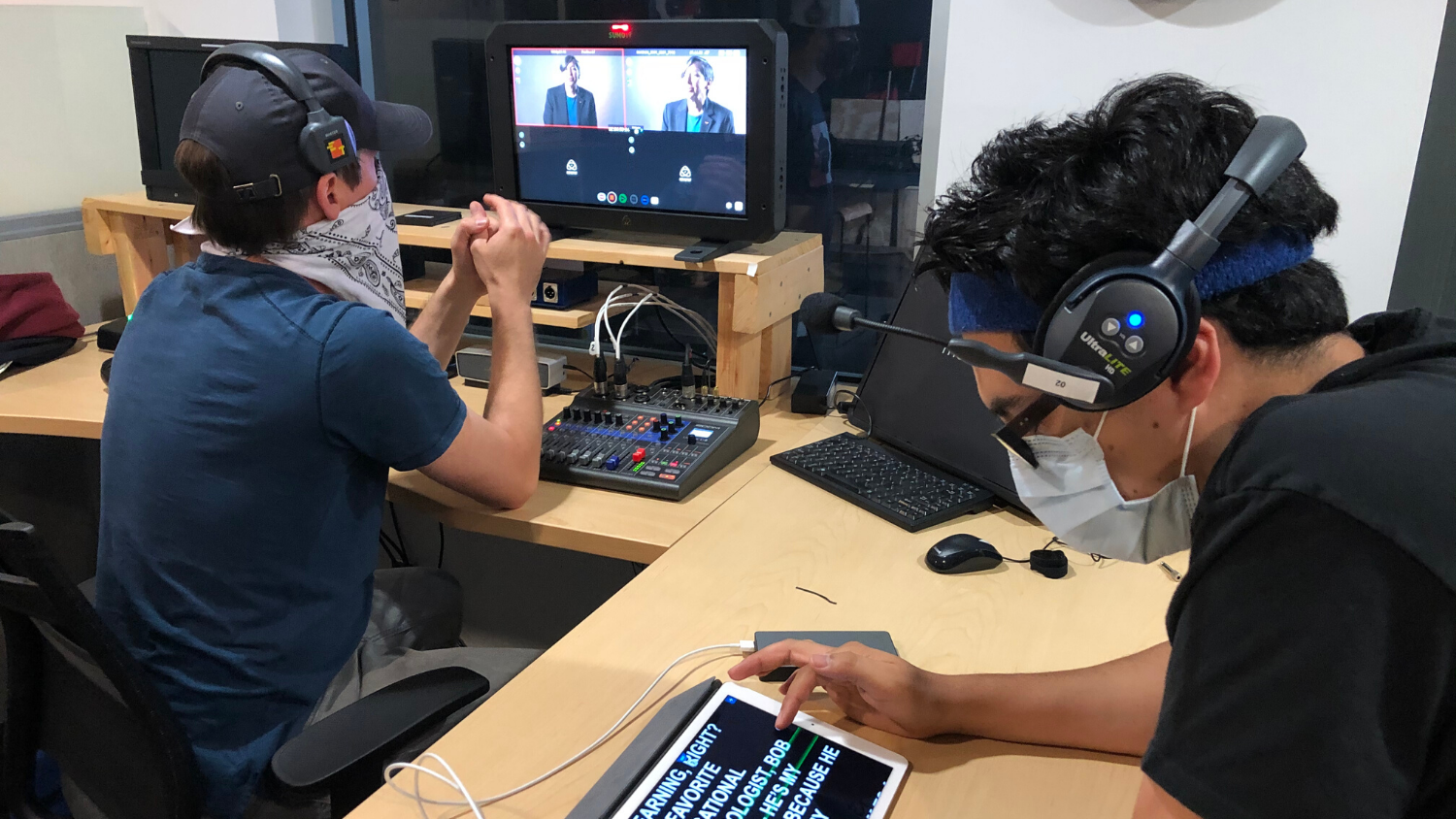 Two men wearing masks and headsets watch recorded footage at a desk in the instructional media production control room.
