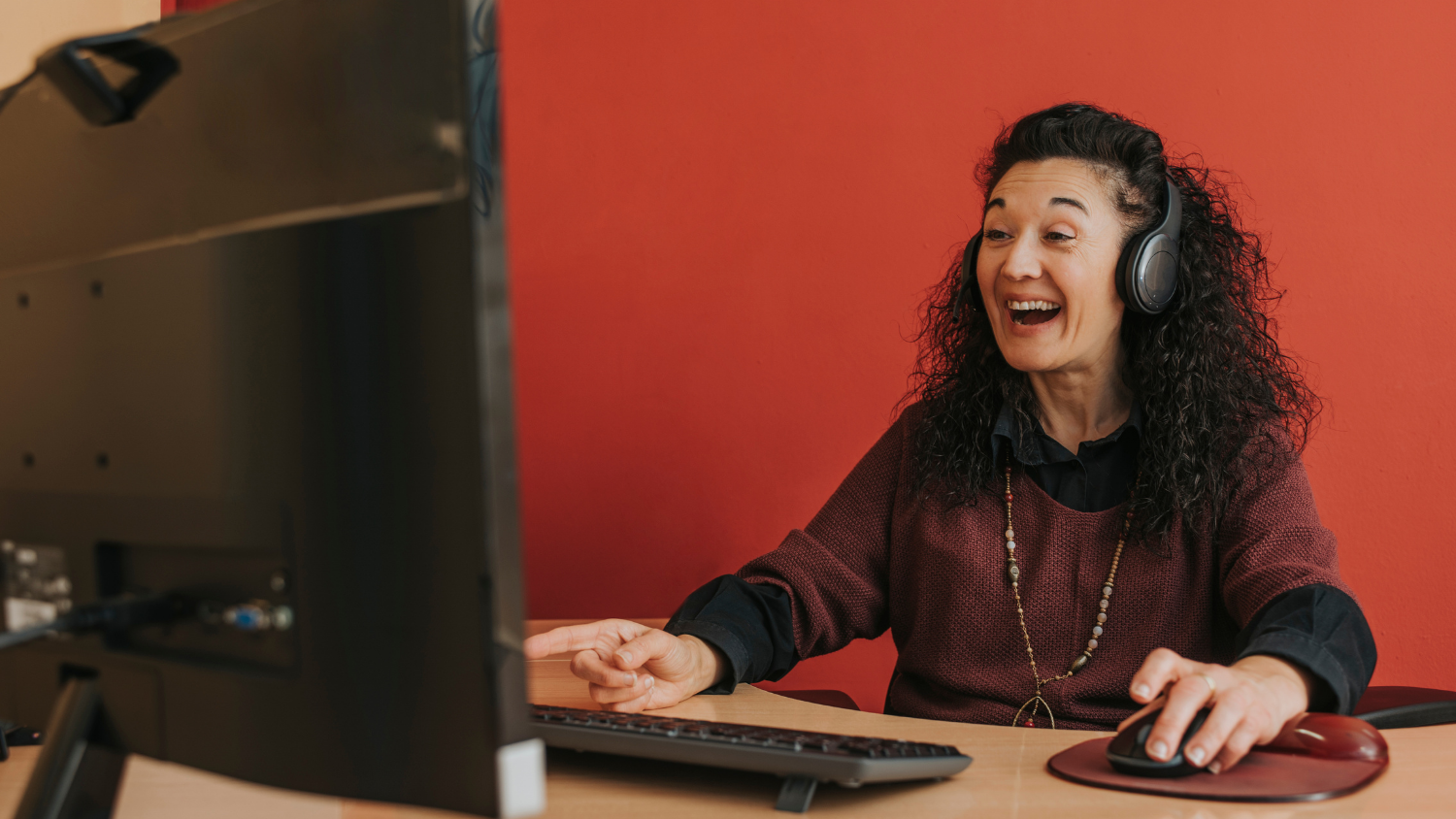 Woman sits in front of computer screen at desk and smiles/laughs.