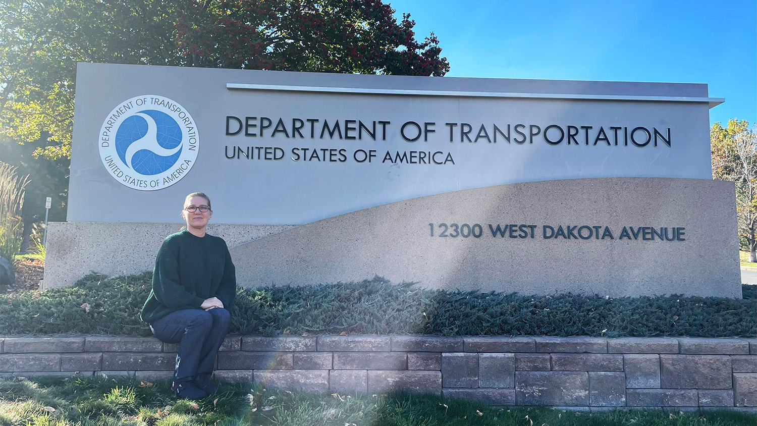 NC State graduate Chelly Sundermeyer sitting in front of a sign outside the Federal Highway Administration Colorado Division