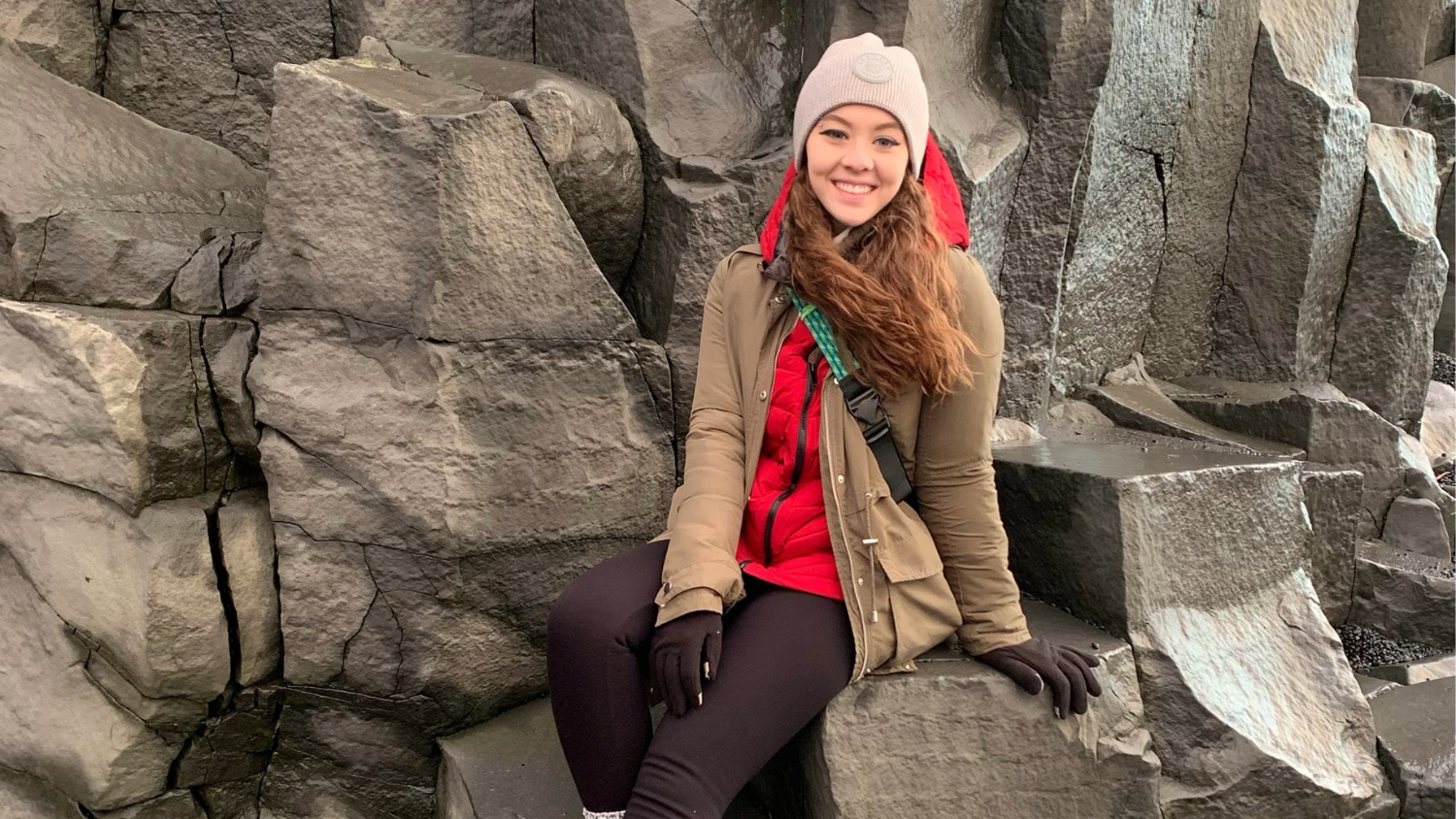 A woman sitting in front of a large rock wall, wearing cold weather gear.