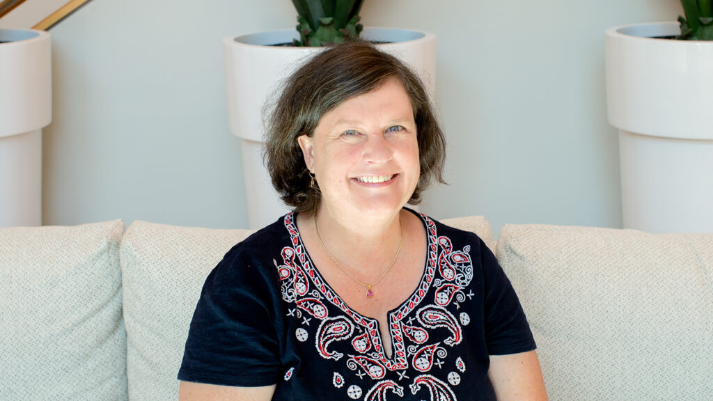 A smiling white woman wearing a black shirt with a colorful decorative design sitting on a couch.