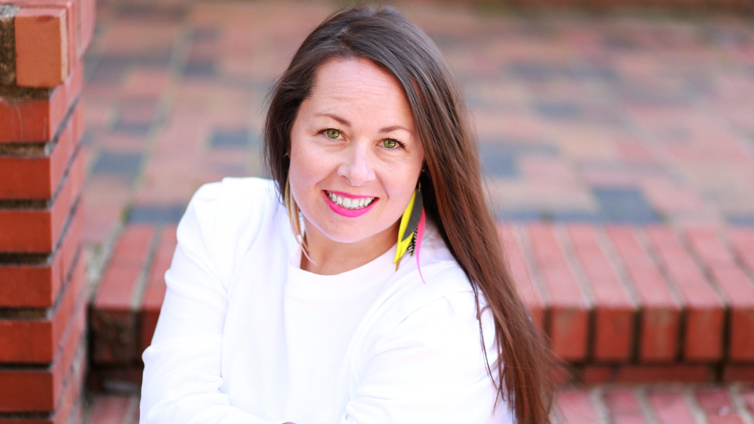 A white woman with long brown hair posing outside on brick steps wearing a white top and colorful feather earrings.