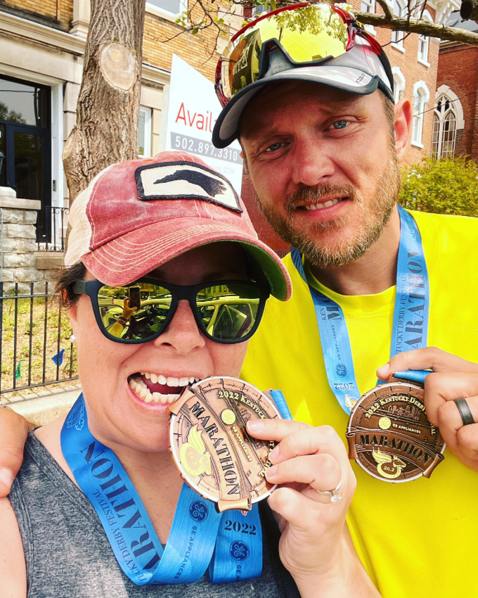 A man and a woman posing outdoors with medals from a road race