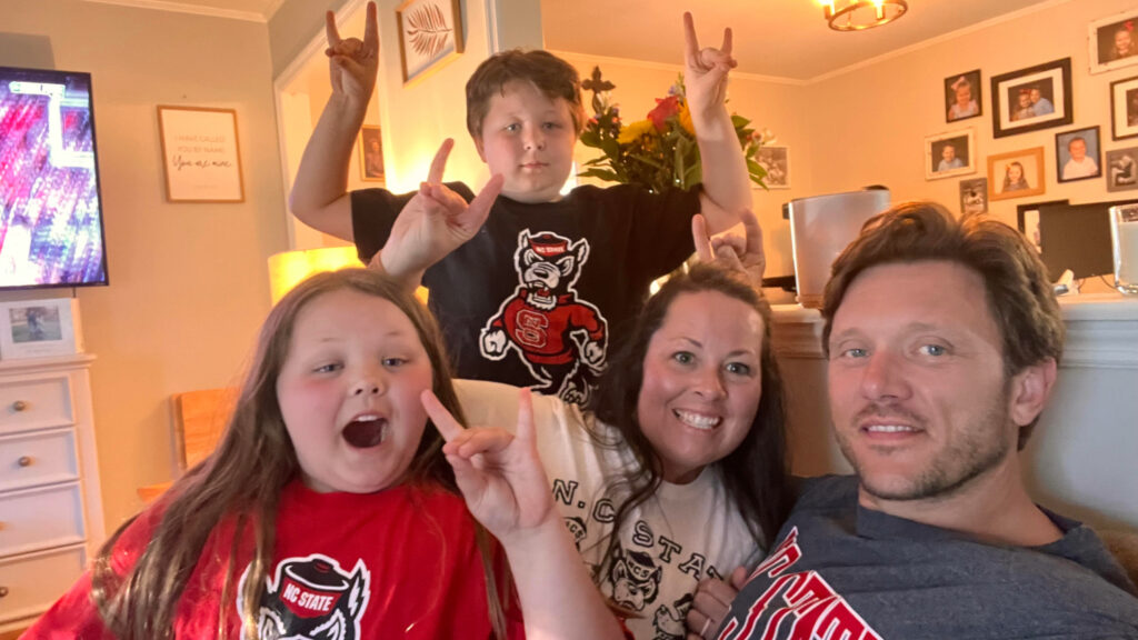 A family wearing NC State clothing cheer with a basketball game on in the background.