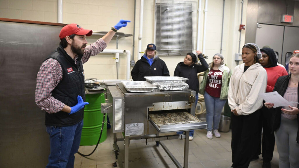 A man standing in front of several students talking. They are in a factory or processing plant and are wearing personal protective gear.