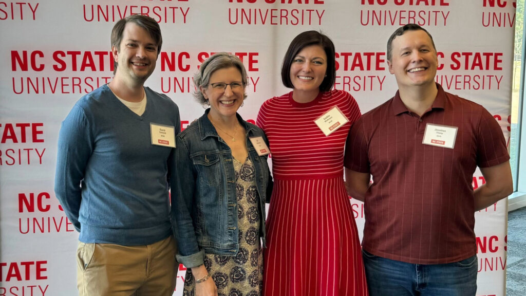 Four people posing in front of a banner reading "NC State University"