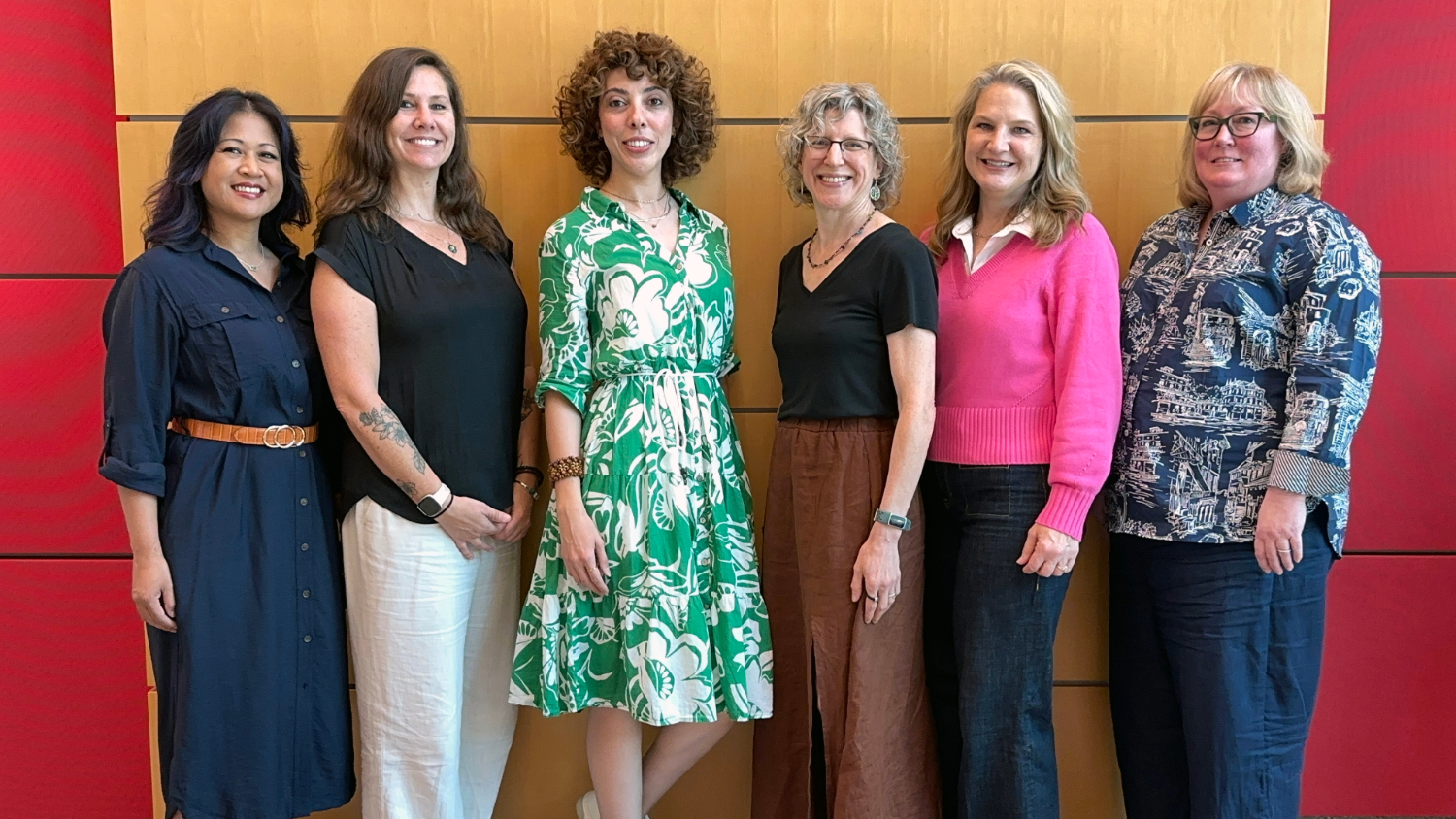 Six women posing indoors in front of a neutral background