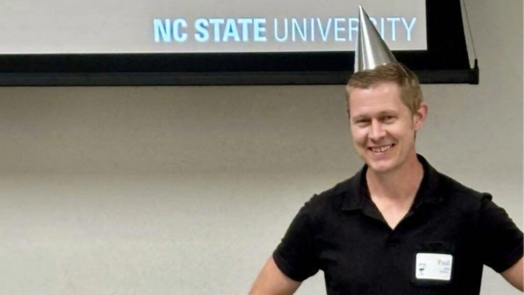 A man standing in a classroom, smiling, with a party hat on