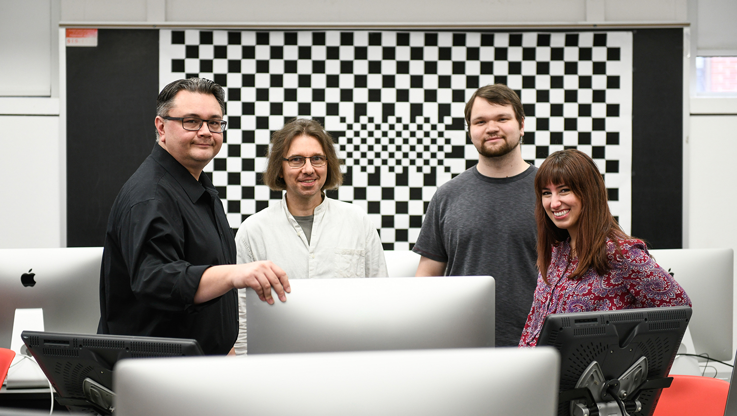 Decorative photo of four people standing around several laptops